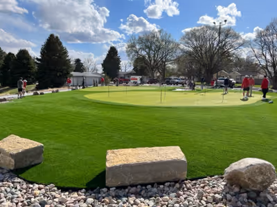 Golf putting green with several people practicing putting under a blue sky with clouds, surrounded by trees and rocks.