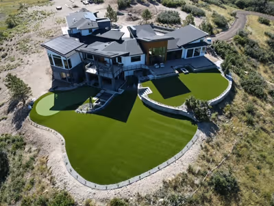 Aerial view of a modern luxury home with expansive artificial turf lawn and putting greens surrounded by natural terrain.