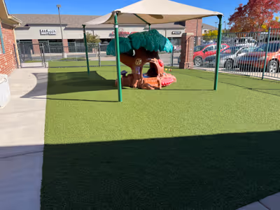 Playground area with artificial turf, a shaded treehouse play structure, and a surrounding fence with parked cars outside.
