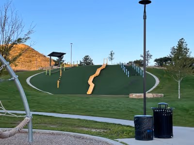 Public park playground with a curved green hill, slides, climbing structures, and a shaded bench area under clear blue sky.