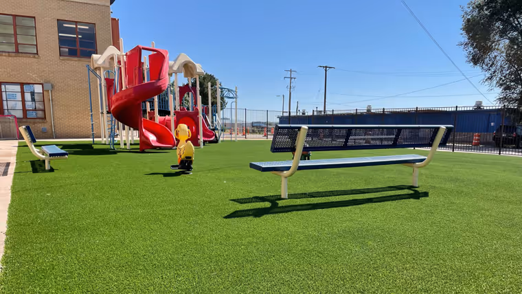 Playground area with bright artificial turf, red spiral slide, spring rider toy, and two metal benches under clear blue sky.