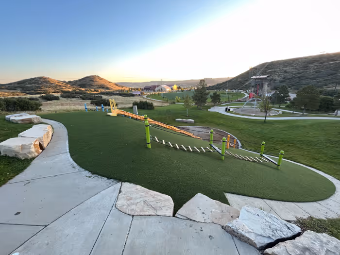 Playground area with bright artificial turf, red spiral slide, spring rider toy, and two metal benches under clear blue sky.