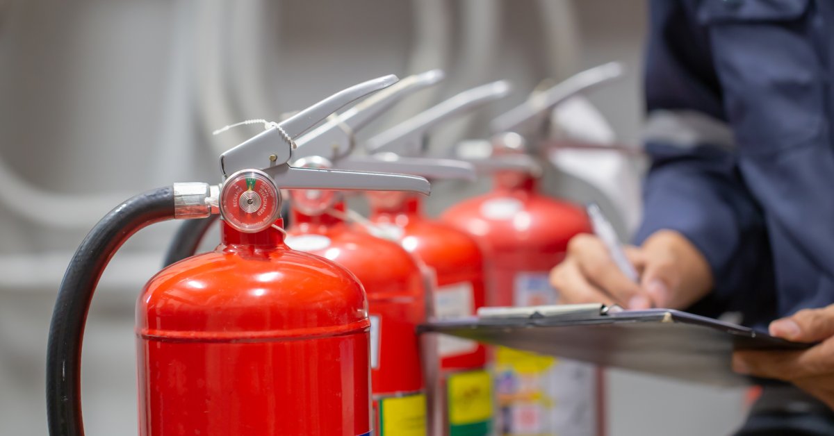 Four fire extinguishers stand upright in a row. A person stands by the extinguishers and takes notes on a clipboard.