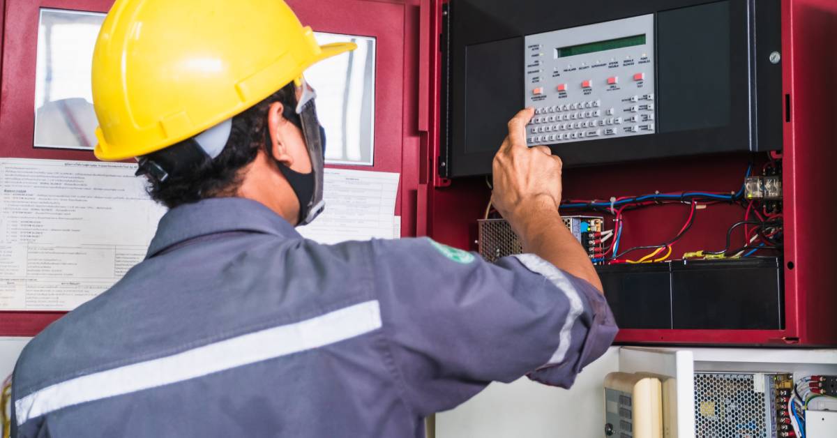 A technician in a yellow hard hat standing in front of an open fire alarm control panel and pressing a button.