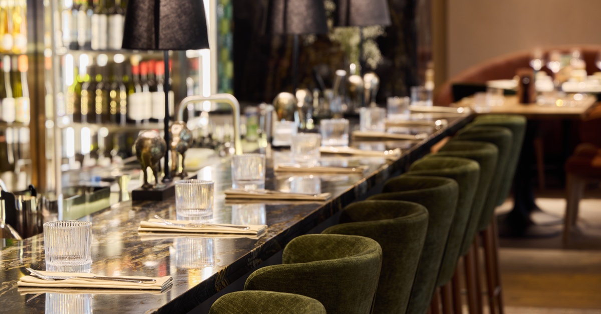 A bar area inside a restaurant features small green chairs on the edge of the counter and various bottles of alcohol.