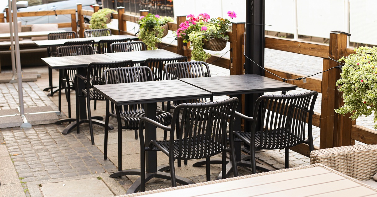 A restaurant's outdoor area features multiple tables and chairs. A blue van sits parked in the background.