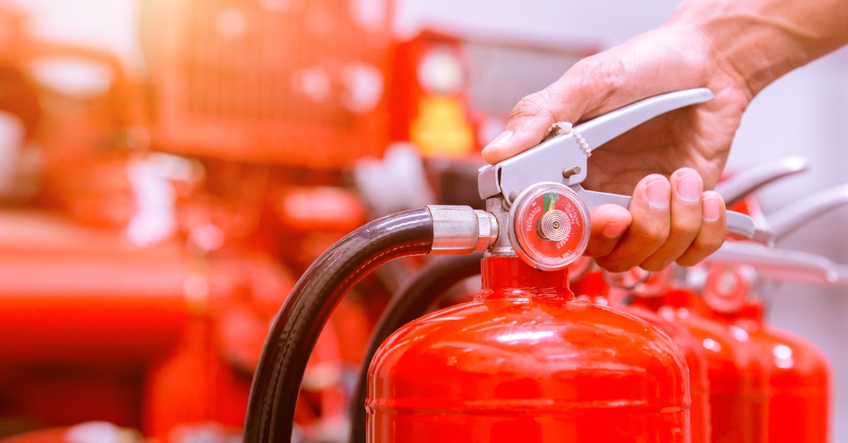 A hand holds onto the silver lever of a red fire extinguisher. It sits next to other fire extinguishers on a floor.