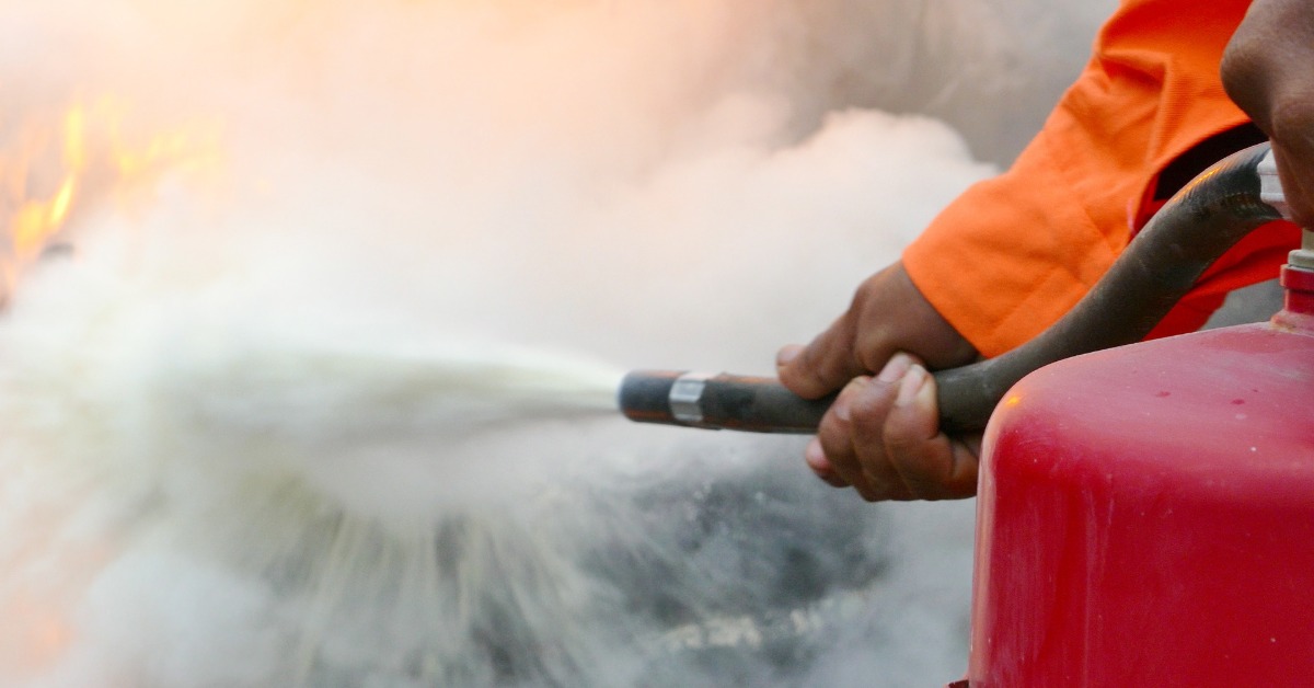 A person holds onto the nozzle and the top of a fire extinguisher and sprays a fire with white powder.