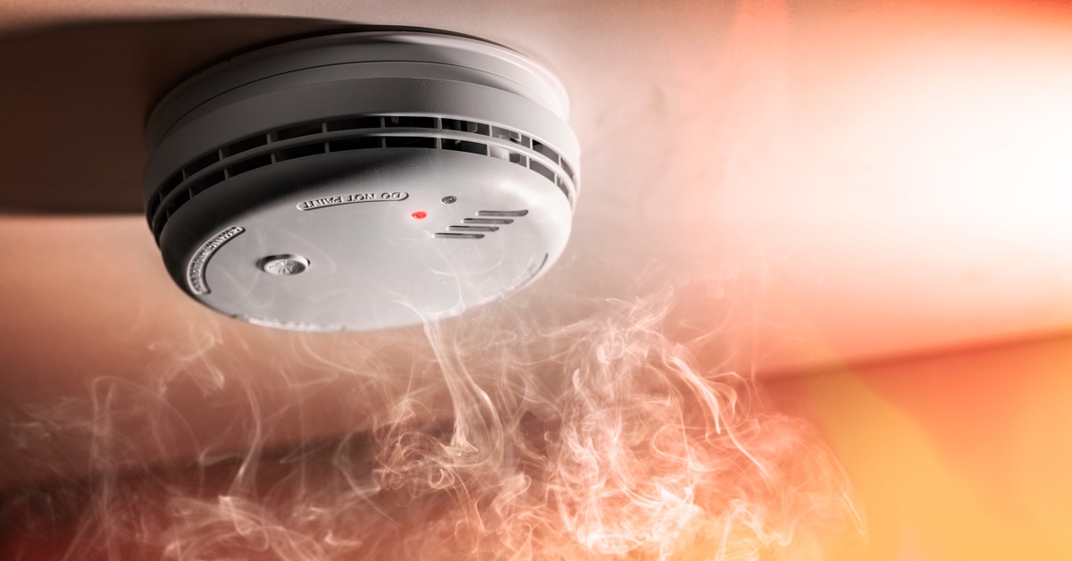 Smoke rises to the ceiling where a smoke detector sits. A small red light shines on the smoke detector.