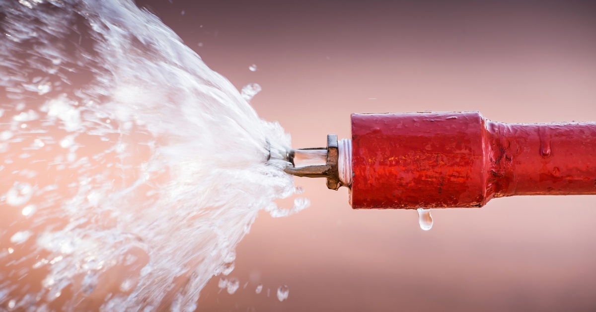 A red pipe with a metal fire sprinkler head sprays crystal clear water. It sits in front of a tan background.
