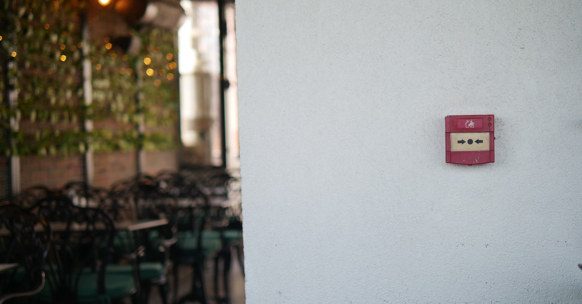 A fire alarm sits on a white wall in a restaurant. Several empty chairs and tables sit in the background.