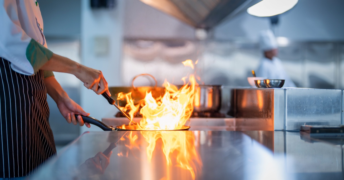 A chef wearing a black apron cooks a flambe in a pan in a restaurant's kitchen. The flame in the pan rises high.