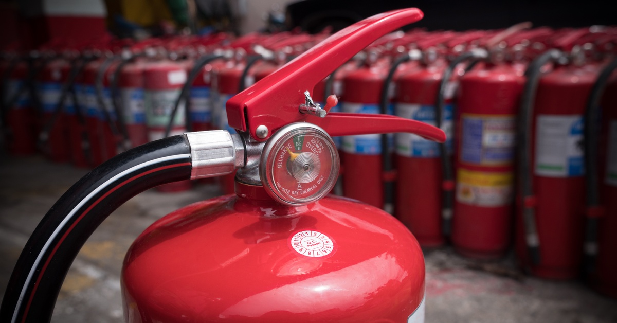 Close-up of a fire extinguisher and its gauge. It sits on the floor of a room in front of a row of other extinguishers.