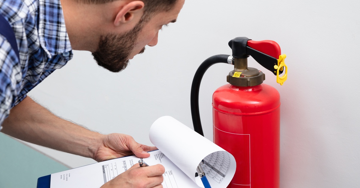 A man with a clipboard makes notes as he inspects a fire extinguisher. It sits on the floor of a building against a wall.