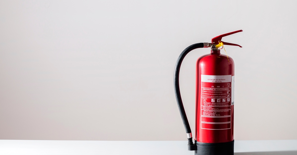 A fire extinguisher sits on a white counter in front of a white wall. The extinguisher has information printed on its side.