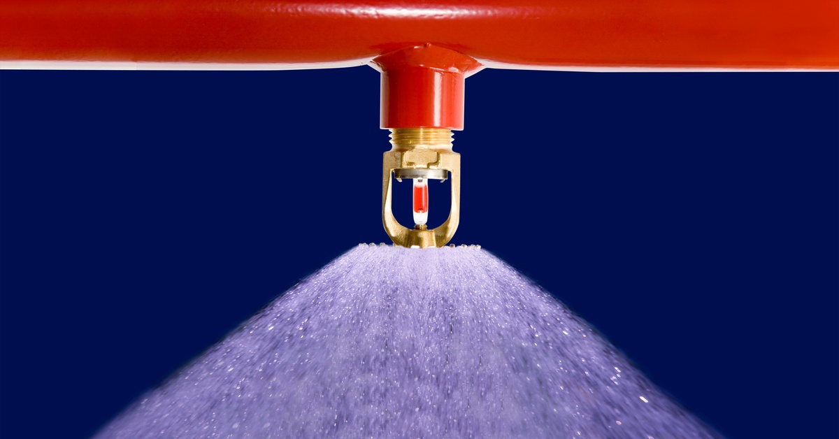 A red pipe features a fire sprinkler head that sprays clear water. It is in front of a blue background.
