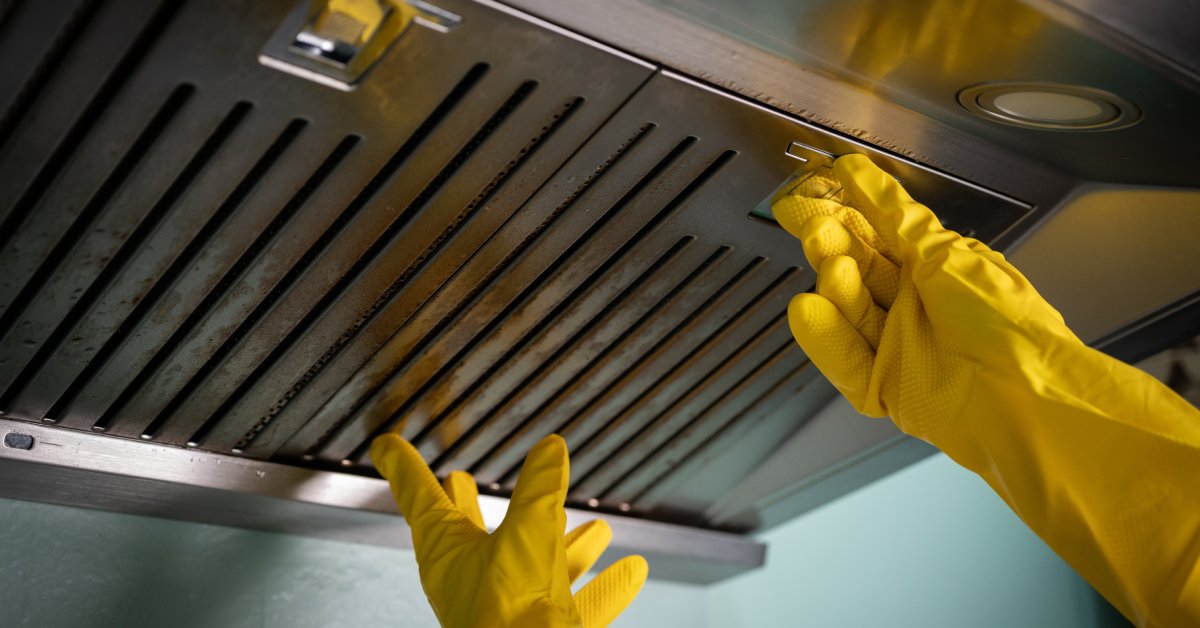 A pair of hands wearing yellow gloves tries to remove a filter from a kitchen oven hood to clean it.