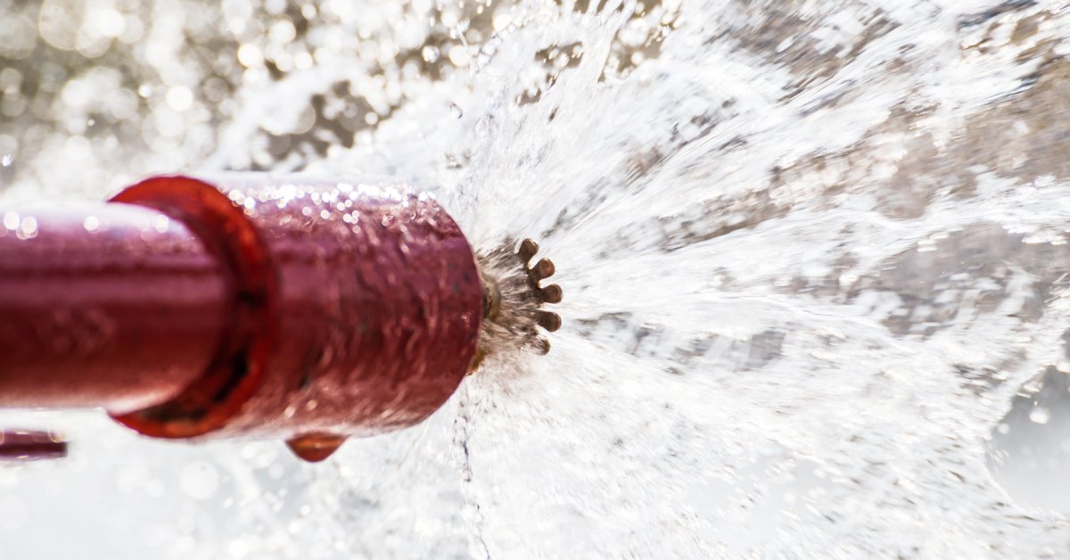 A sprinkler system spraying out a healthy stream of clear water from its nozzle. The sprinkler's pipe is red.