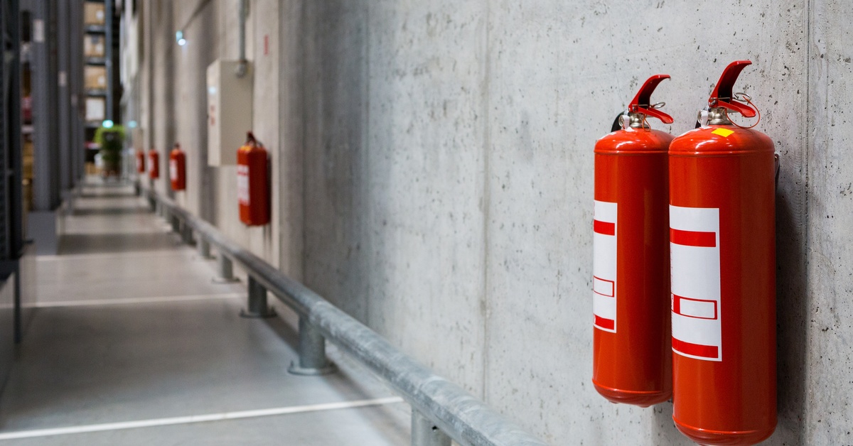Multiple fire extinguishers hang in pairs on the walls of a warehouse. They are across from storage racks.