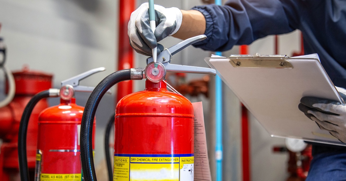 Close-up of a person's hands as they inspect a fire extinguisher. They wear gloves and hold a pen and a clipboard.