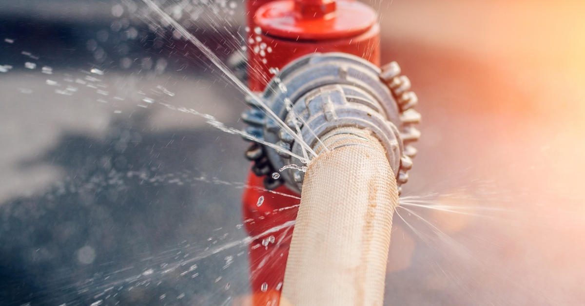 Close-up of a large fire hose attached to a fire hydrant. The hose leaks and sends out trickles of water.
