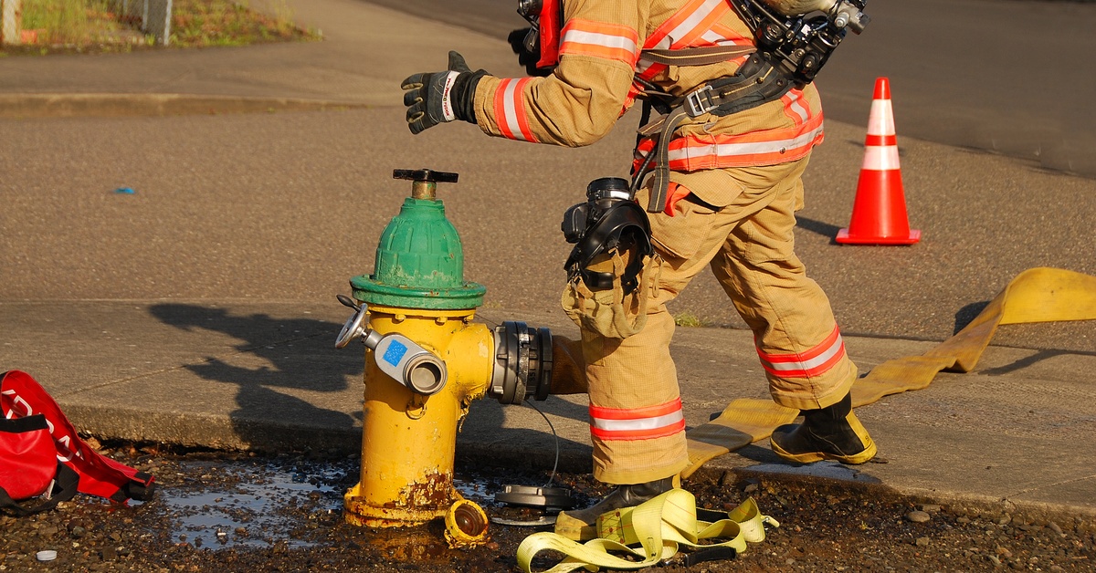 A fireman wearing his gear is attaching a hose to a yellow fire hydrant. An orange parking cone stands nearby.
