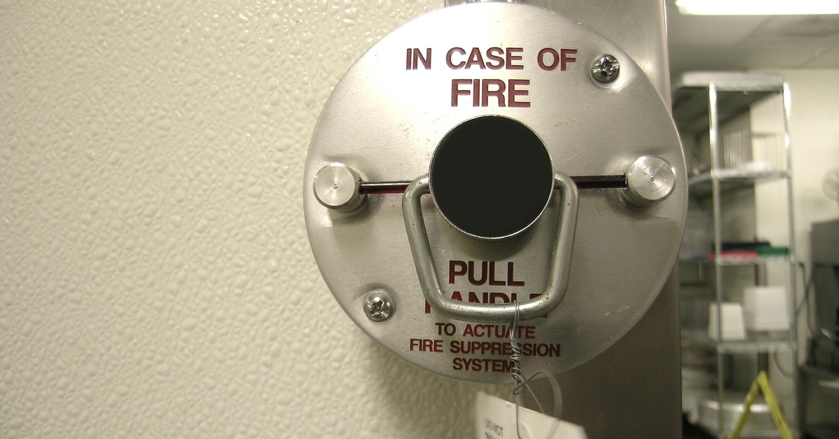 A silver fire suppression system handle on a restaurant kitchen wall reads