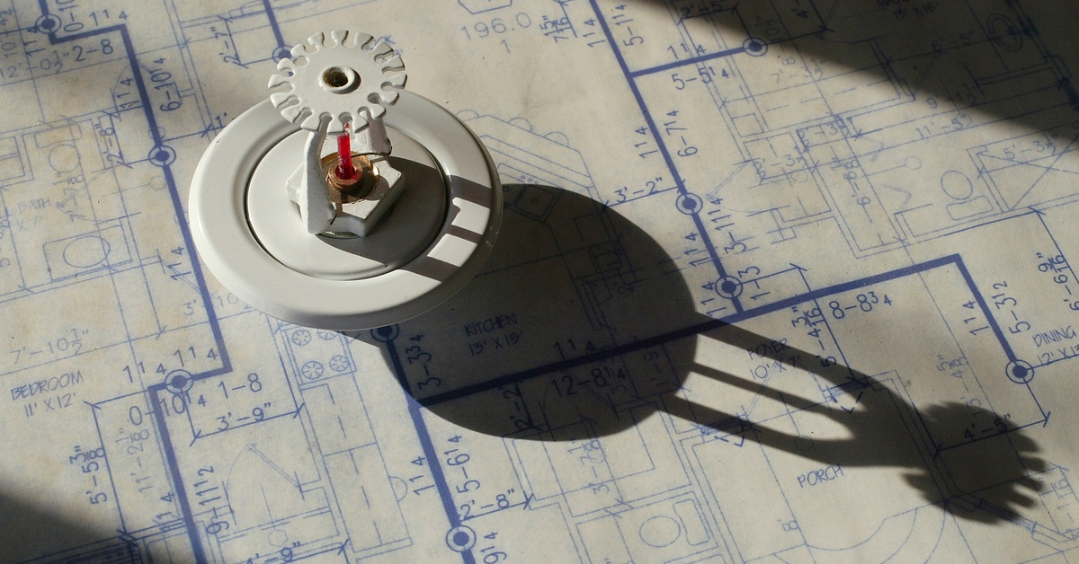 Light shining on a sprinkler head sitting on top of a building blueprint. The sprinkler head's shadow stretches across the page.
