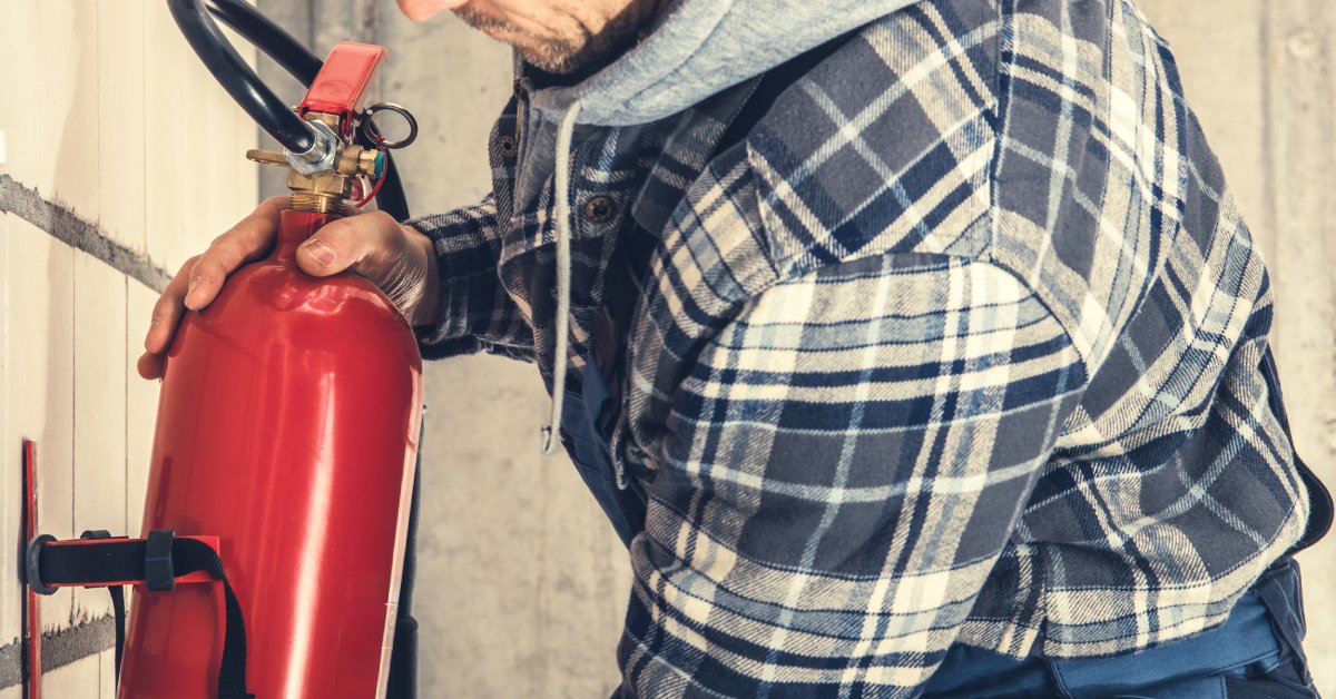 A worker wearing dark safety glasses while mounting a fire extinguisher to a tiled wall. He holds the top and bottom securely.