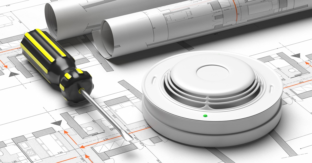 A smoke detector, a screwdriver, and two rolled-up blueprints lie on top of a blueprint showing room layouts.