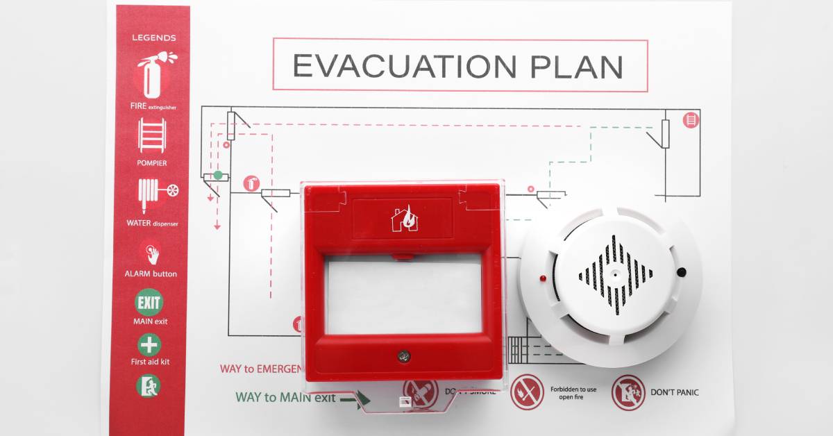 A smoke detector and manual fire alarm call point box sitting on top of a fire evacuation plan. The plan shows exit routes.