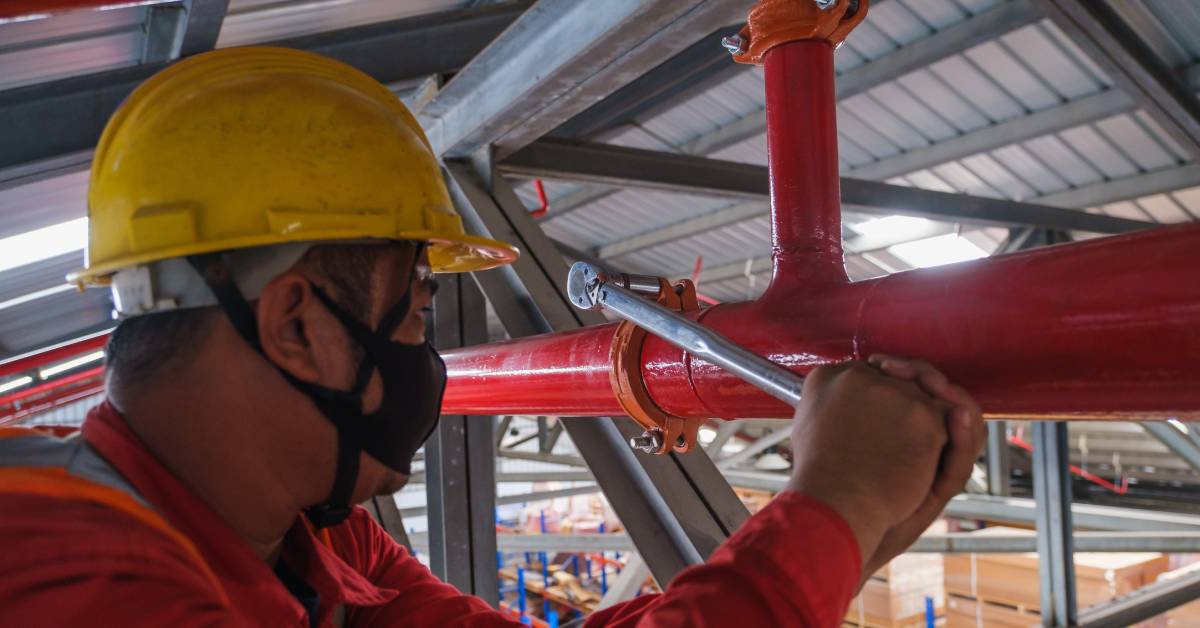 A technician adjusts a component on a red water pipe with a socket wrench. He works near the ceiling of a warehouse facility.