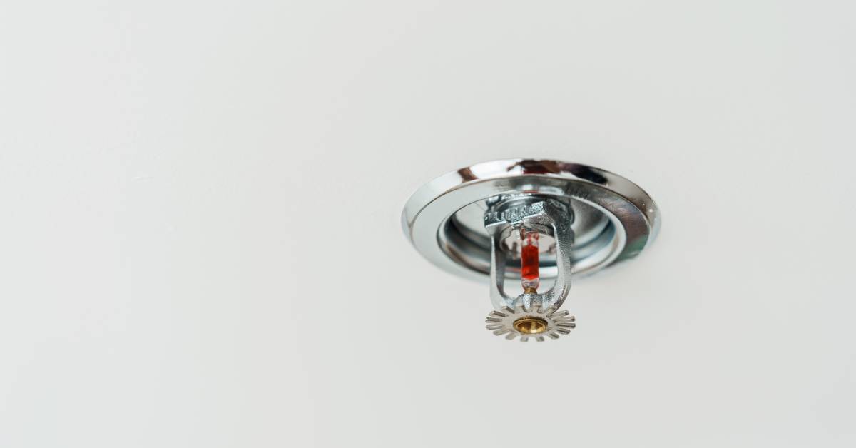  A shiny fire sprinkler head protrudes from a smooth white ceiling. The sprinkler head's glass bulb has red liquid inside.