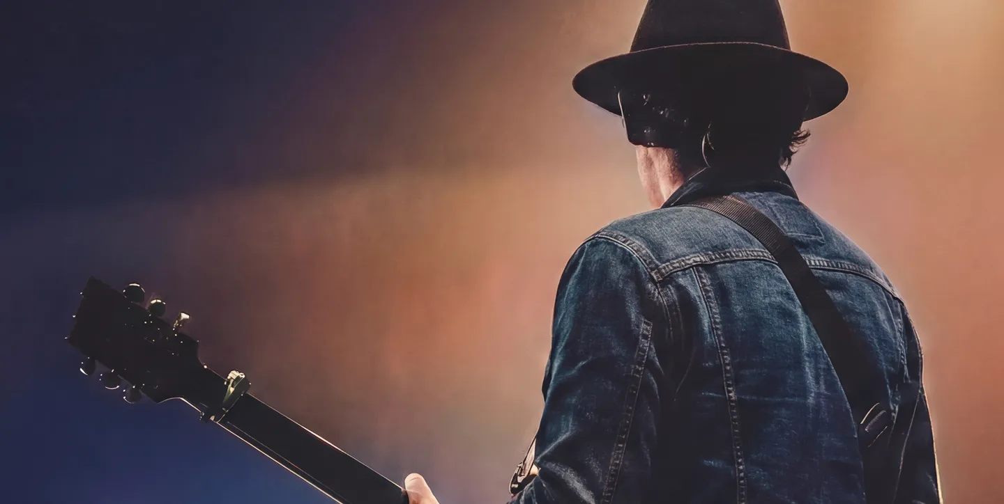 Musician wearing a black hat and denim jacket playing guitar on stage with warm lighting.