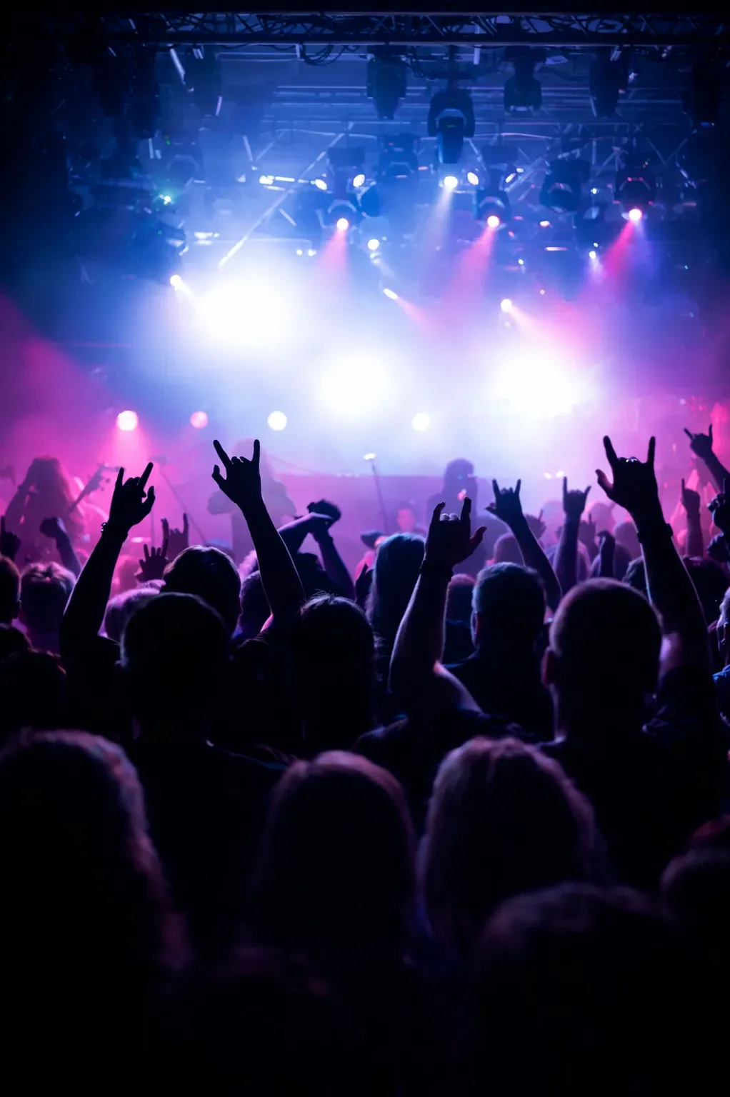 Crowd at a concert with raised hands making the rock and roll gesture under colorful stage lights.