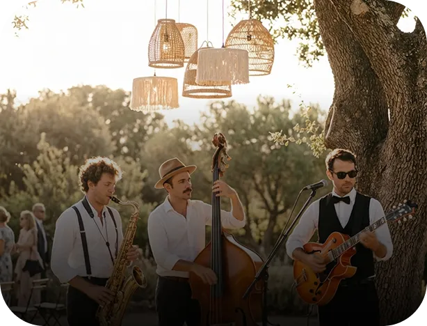 Three musicians playing saxophone, double bass, and guitar outdoors under hanging wicker lamps.