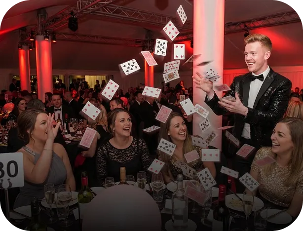 A magician in a black tuxedo performing a card trick with cards flying around a smiling table of four women in a festive event setting.