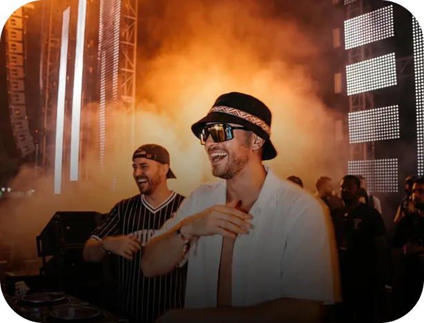 Two men smiling and enjoying music on a stage with bright lights and smoke effects in the background.
