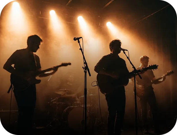Silhouettes of four musicians playing guitars and drums on stage under warm spotlights and smoky atmosphere.