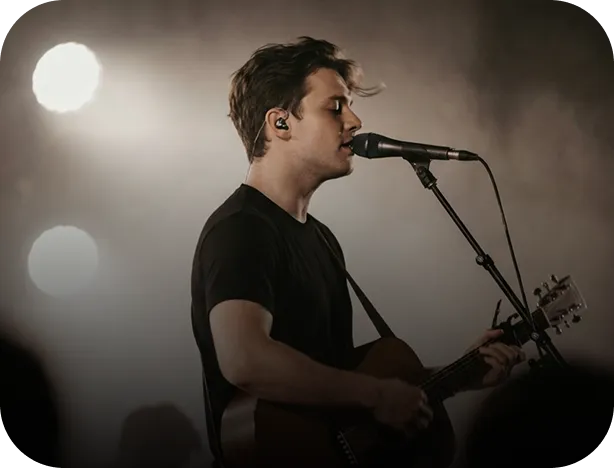 Male musician singing into a microphone while playing an acoustic guitar on stage with backlighting.
