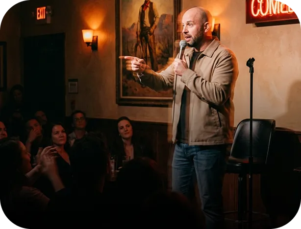 Comedian in brown jacket and jeans performing stand-up in front of an audience at a comedy club.