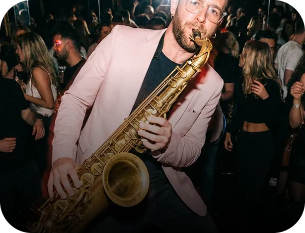 Man in a pink blazer playing a saxophone at a crowded indoor event or party.