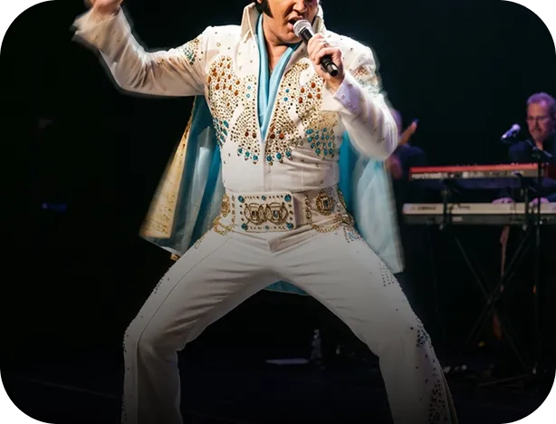 Performer in a white embellished jumpsuit with a microphone mid-performance on stage with keyboard player in background.