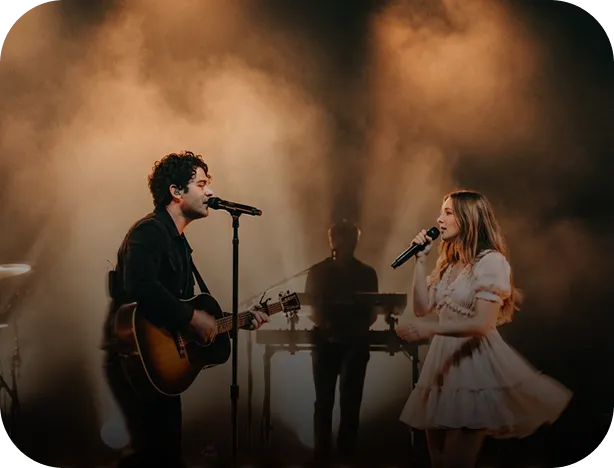 Male musician playing guitar and singing into microphone facing female singer in white dress holding microphone on stage with dramatic lighting and smoke.
