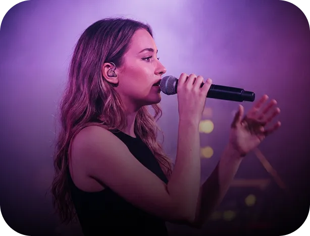 Woman with long hair singing into a microphone on a purple-lit stage.