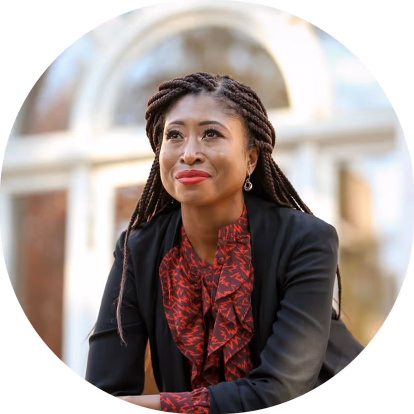 Portrait of a woman with long braided hair wearing a black blazer and red patterned blouse, looking slightly upward.