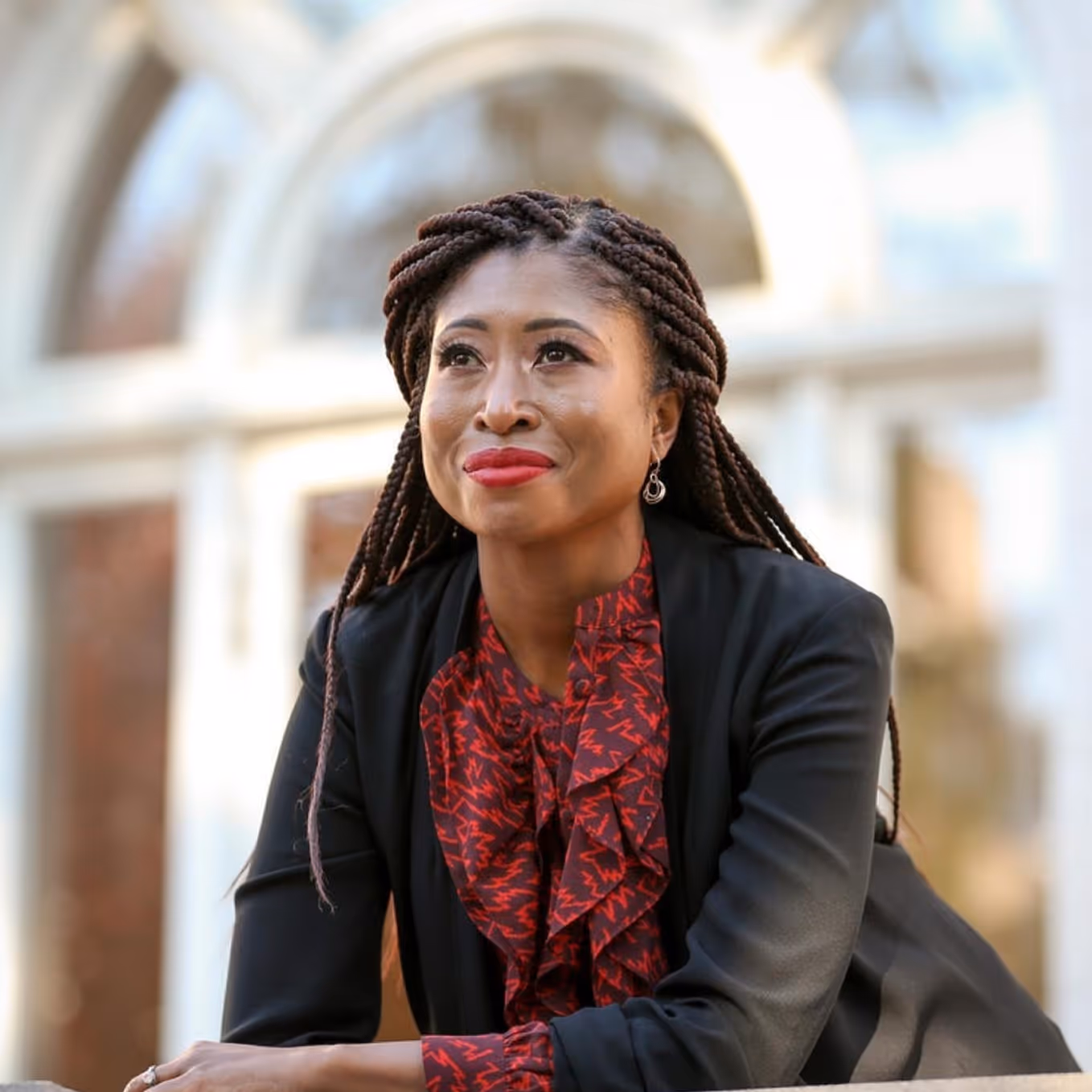 Smiling woman with braided hair wearing a black blazer and red patterned blouse sitting outdoors in front of a blurred building.