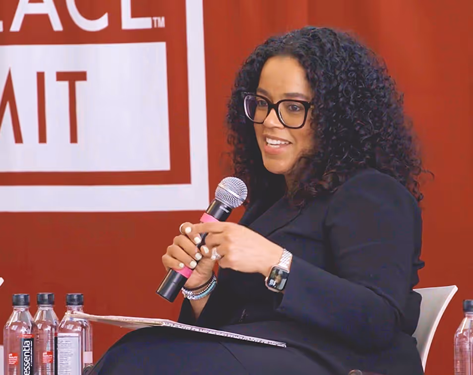 Woman with curly hair and glasses speaking into a microphone while seated, with water bottles and a sign in the background.