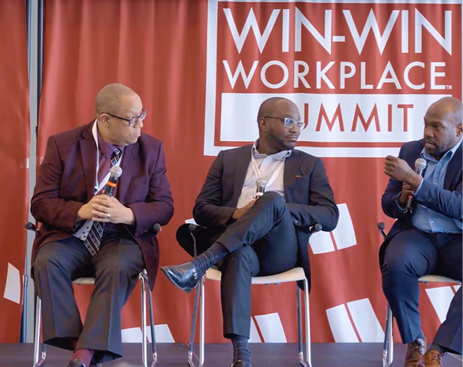 Three men in suits seated on stage holding microphones, participating in a panel discussion at the Win-Win Workplace Summit.