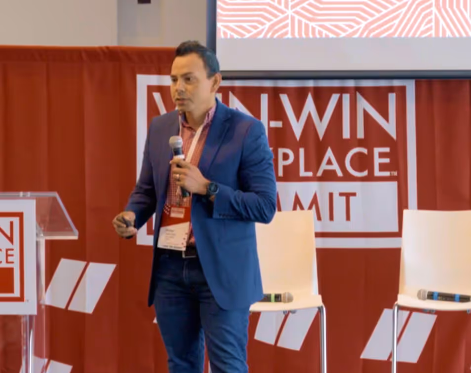 Man in blue blazer holding a microphone, speaking at Win-Win Workplace Summit with chairs and a screen in the background.